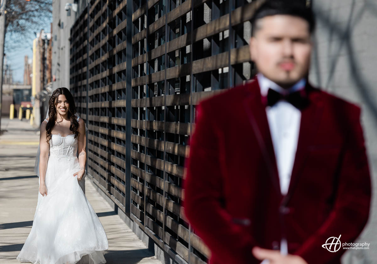 A groom dressed in a classic black suit stands with his back facing the camera, waiting for his bride for their first look. In the background, the bride is walking towards him, wearing a stunning white wedding dress with a long train. The groom looks anxious and excited, while the bride appears calm and composed. The scene is set against a beautiful outdoor backdrop, with lush greenery and trees surrounding the couple. The anticipation and love between the couple is palpable in this moment, and it is clear that their wedding day is going to be a special and unforgettable event.