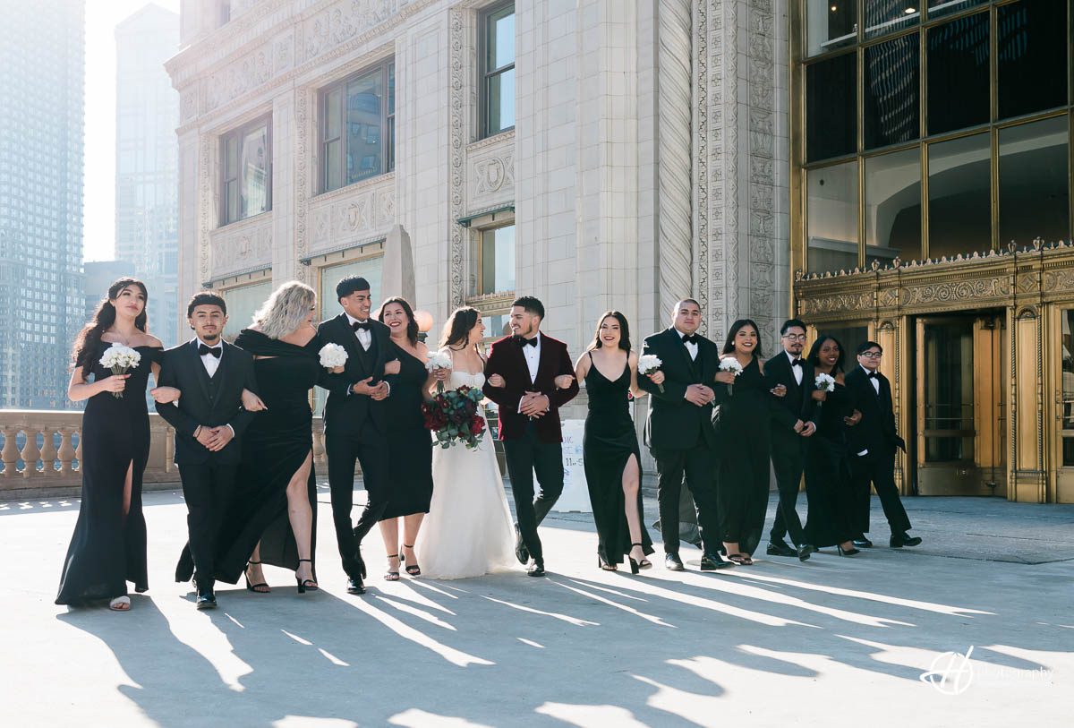 A group of bridesmaids and groomsmen are seen walking and talking along the Chicago River Esplanade, dressed in stylish and coordinated wedding attire. The background features a stunning view of the river, with skyscrapers and city buildings in the distance. The bridal party appears to be enjoying each other's company and the picturesque scenery as they make their way to the wedding venue.