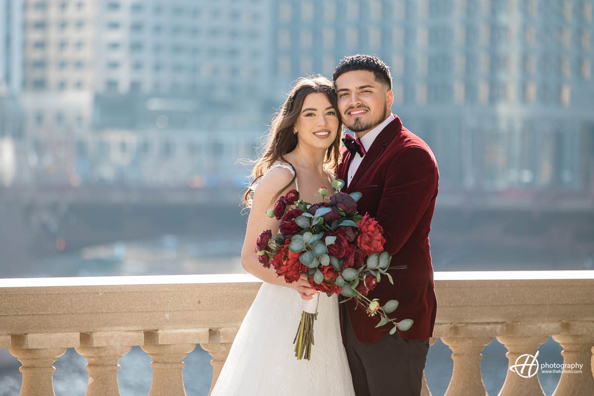 In the portrait, Samuel and Faith are standing close to each other, with the beautiful Chicago skyline visible in the background. Both are dressed elegantly, with Faith wearing a stunning white wedding gown and Samuel in a sharp red suit. They are looking lovingly into each other's eyes, with a sense of joy and happiness emanating from their expressions. The towering skyscrapers of Chicago provide a breathtaking backdrop, creating a romantic and picturesque scene