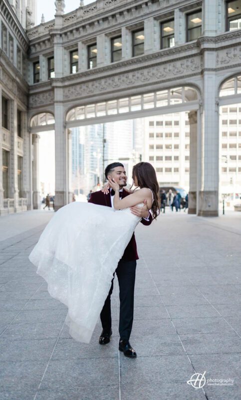 A romantic image captures a groom holding his bride in his arms in front of the iconic Wrigley Building. The groom is dressed in a stylish suit, while the bride is wearing a beautiful white gown. They both have big smiles on their faces as they look into each other's eyes. The Wrigley Building, a historic skyscraper, serves as a breathtaking backdrop, with its distinctive clock tower and ornate architectural details. The sun is setting in the background, casting a warm glow on the scene, creating a truly magical moment.