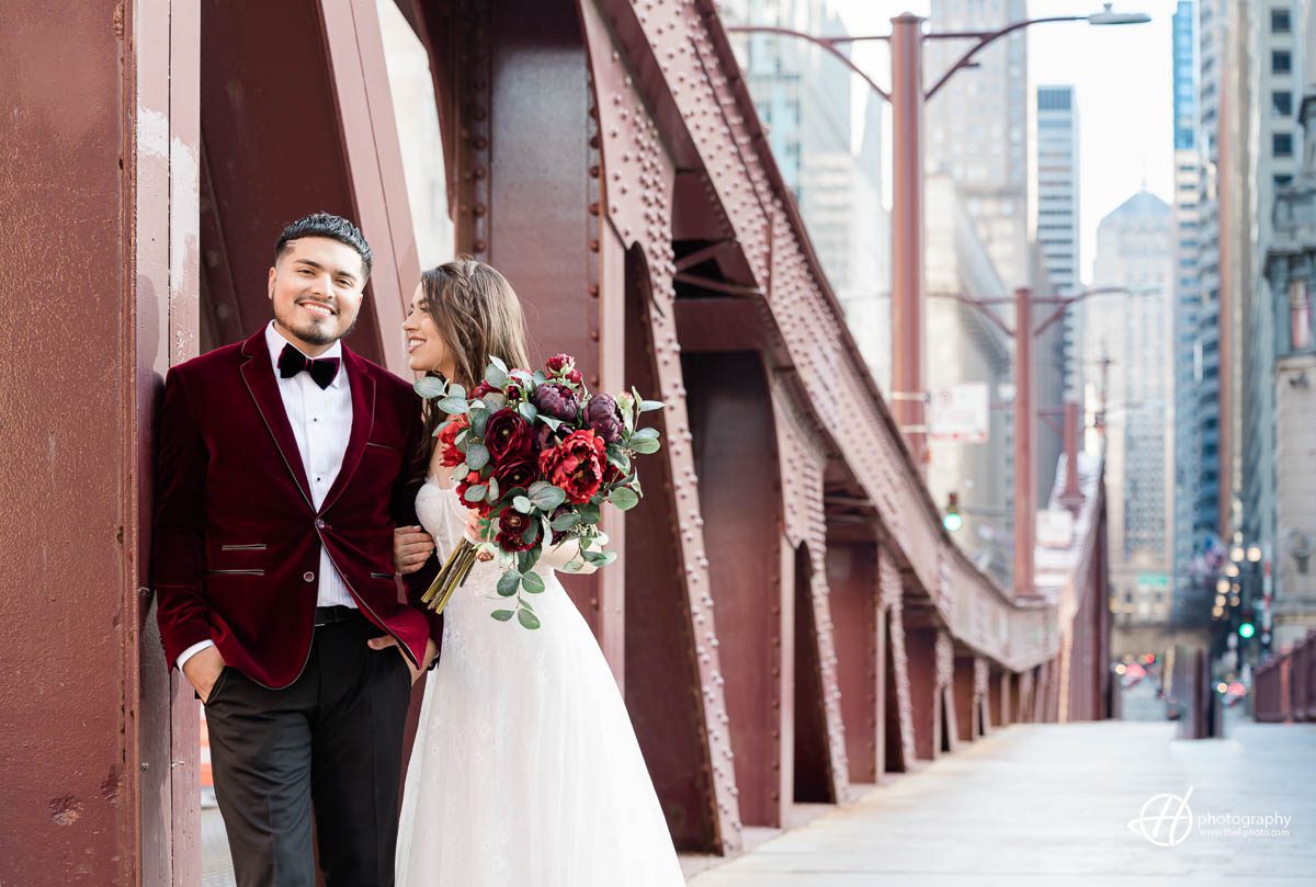 Samuel and Faith pose for a photo on LaSalle Bridge in Chicago, with the Chicago River flowing underneath. They stand side by side, smiling at the camera. The bridge offers a picturesque view of the city's skyline in the background, with tall buildings towering over the river