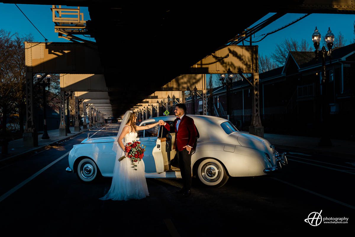 bride and groom are posing in front of a Rolls Royce car, parked on a street under a train track in Chicago. The bride is wearing a white wedding gown and holding a red bouquet of flowers, while the groom is dressed in a red suit and tie. The car is white and polished, with a luxurious leather interior visible through the open door. The train track overhead is elevated and has a metal structure, casting a shadow on the couple and the car. The background shows a city street with buildings and cars parked along the side. The couple looks happy and stylish, ready for their wedding day