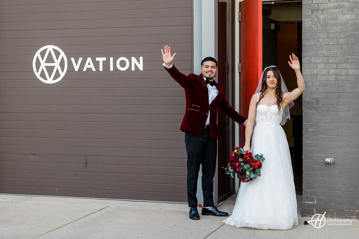 Samuel and Faith, are standing outside Ovation wedding venue, waving at the camera. Samuel is wearing a red suit and Faith is dressed in a white wedding gown. The building behind them is a modern, two-story brick structure with large windows and a sign that reads "Ovation". The surrounding area is well-maintained, with manicured lawns and bushes in the background. The couple looks happy and excited, perhaps ready to start their wedding reception inside the venue. 