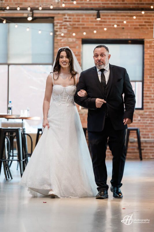 An emotional scene is captured in this image, as a bride and her father are shown walking down the aisle at Ovation wedding venue. The bride is wearing a beautiful, white wedding gown with a long veil, and carrying a bouquet of flowers. Her father is dressed in a black suit and tie, holding onto her arm and guiding her down the aisle towards her future husband. The venue's decor is visible in the background, with chairs lined up on either side of the aisle, flowers and greenery adorning the space. The lighting is soft and warm, adding to the romantic atmosphere. The bride looks happy and nervous, perhaps taking in the moment and anticipating the exchange of vows.