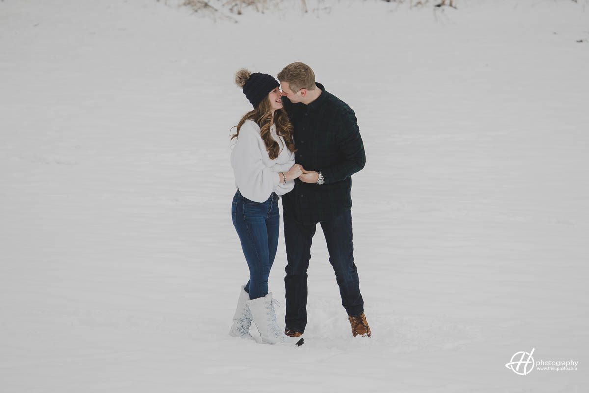 Julia and Jospeh stands on a snowy landscape during their engagement photo shoot, gazing into each other's eyes with smiles on their faces. The snow-covered trees and mountains create a beautiful backdrop, and the couple is bundled up in warm winter clothing.
