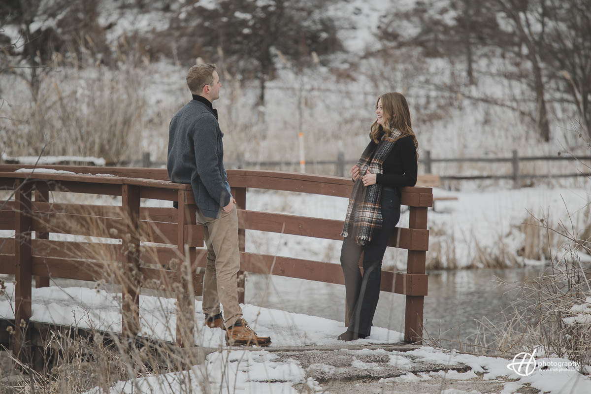 Amidst a winter wonderland, an engaged couple stands on a bridge, chatting and smiling. The snow-covered trees and icy river provide a stunning backdrop for this intimate moment, as they share in the excitement of their upcoming wedding.