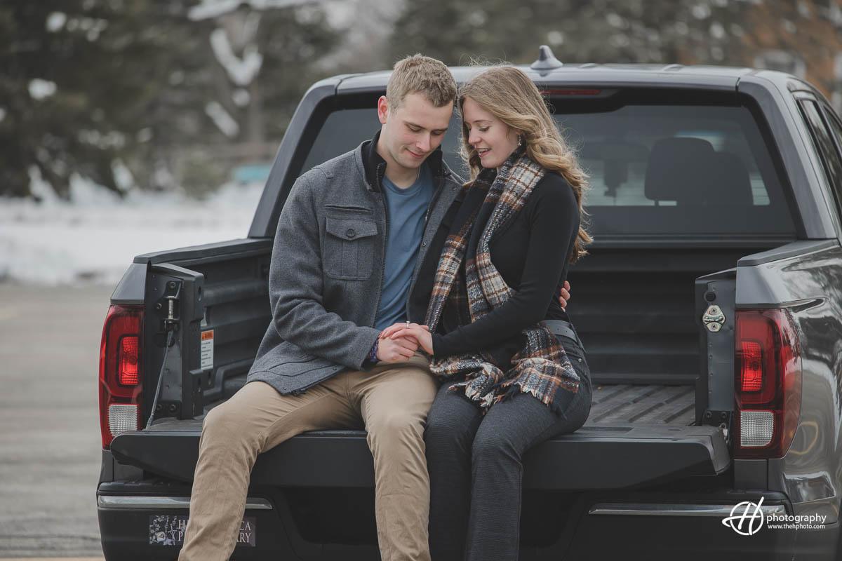ulia and Joseph cozy up in the back of their truck, surrounded by the beauty of a winter wonderland. The snow-covered trees and frozen lake create a serene atmosphere, as they snuggle up together to keep warm. Their love shines bright in this peaceful and romantic moment.